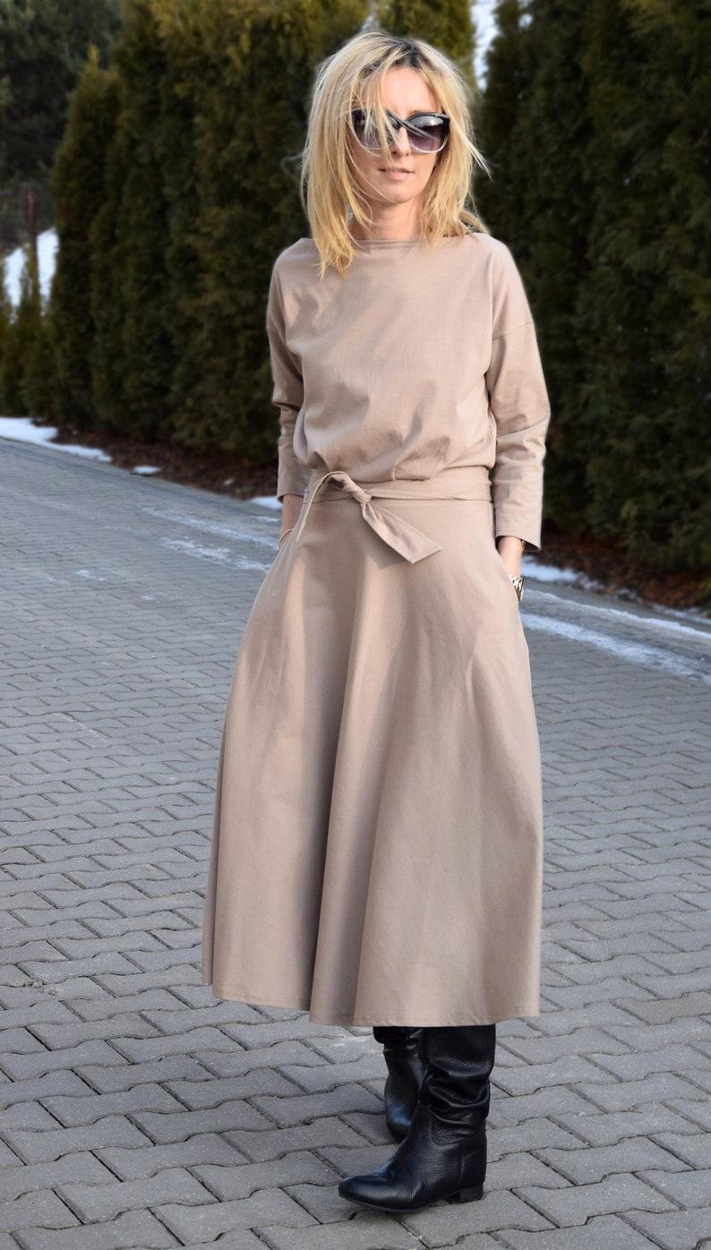 May include: A beige dress with long sleeves and a matching tie belt. The dress has a round neckline and a full skirt. The person is wearing black boots and sunglasses. The background features green trees and a paved walkway.