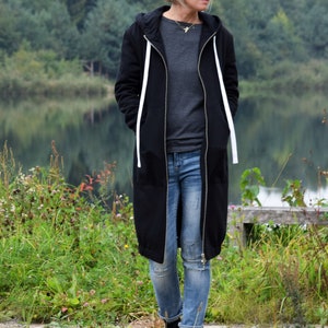 May include: A woman wearing a long black hooded sweatshirt with white drawstrings and blue jeans. She is standing in a grassy area near a body of water.