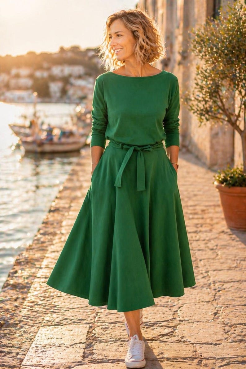 May include: A long-sleeved, emerald green dress with a boat neckline and a matching belt. The dress has a flowing skirt and pockets. The person is wearing white sneakers. The background features a waterfront scene with a boat and a stone walkway.