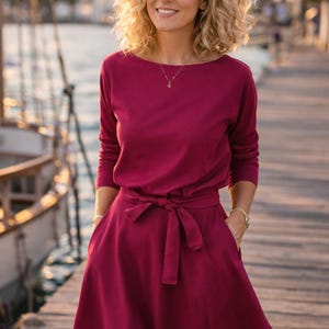 May include: A burgundy dress with long sleeves, a boat neckline, and a tie-waist. The dress features a flowing skirt and pockets. A gold necklace and bracelet are visible. The background shows a wooden dock and water.