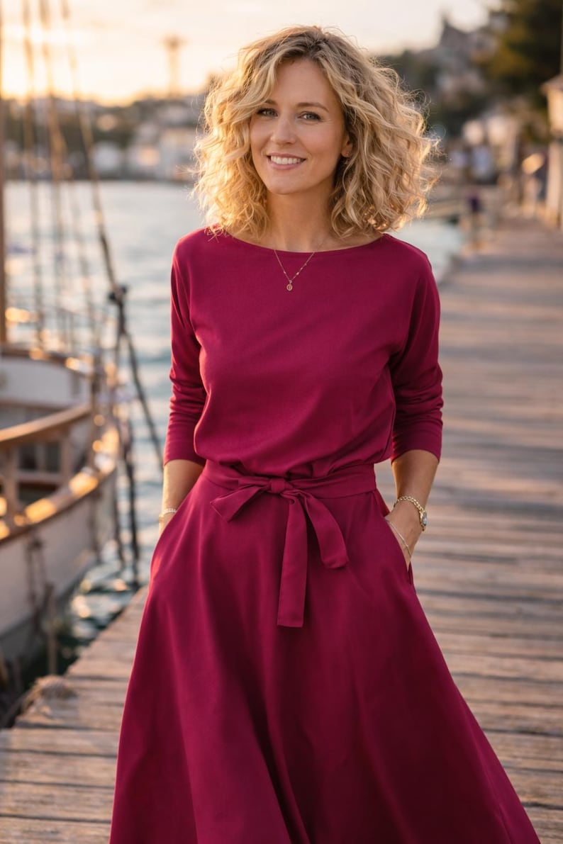 May include: A burgundy dress with long sleeves, a boat neckline, and a tie-waist. The dress features a flowing skirt and pockets. A gold necklace and bracelet are visible. The background shows a wooden dock and water.