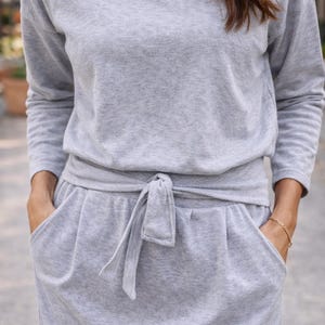 May include: A light gray, two-piece lounge set. The top is a long-sleeved, boat-neck style. The bottoms are a skirt with pockets and a tie-waist detail. The outfit is made of a soft, comfortable material.