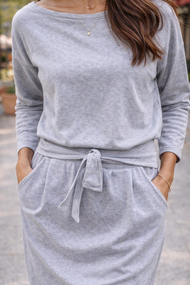 May include: A light gray, two-piece lounge set. The top is a long-sleeved, boat-neck style. The bottoms are a skirt with pockets and a tie-waist detail. The outfit is made of a soft, comfortable material.