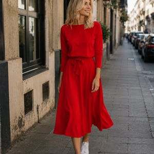 May include: A vibrant red dress with long sleeves and a tie at the waist. The dress is worn with white trainers. The person is walking on a paved path in front of a building with windows.