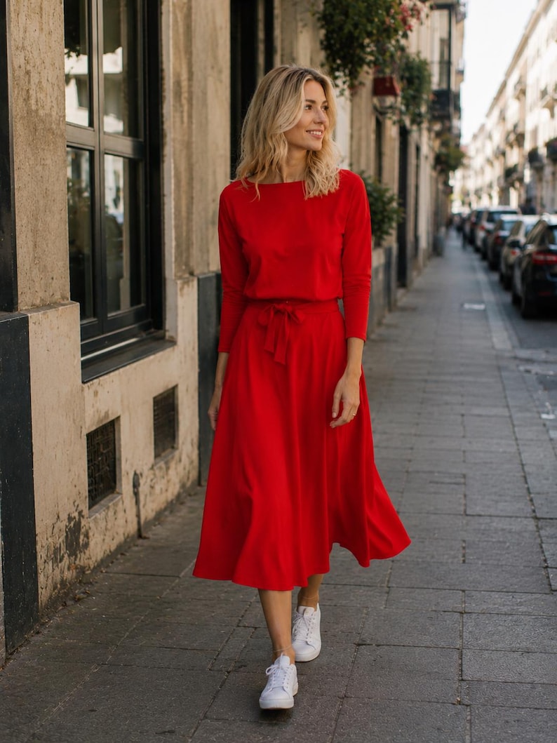 May include: A vibrant red dress with long sleeves and a tie at the waist. The dress is worn with white trainers. The person is walking on a paved path in front of a building with windows.
