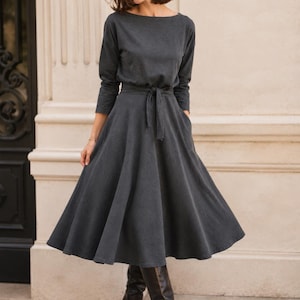 May include: A dark grey dress with long sleeves and a boat neck. The dress has a belted waist and a flared skirt that falls to the knees. The person is wearing black knee-high boots. The dress has pockets.