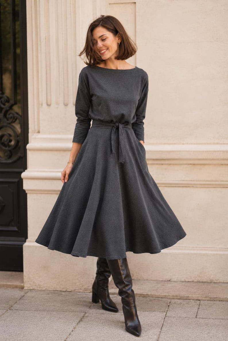 May include: A dark grey dress with long sleeves and a boat neck. The dress has a belted waist and a flared skirt that falls to the knees. The person is wearing black knee-high boots. The dress has pockets.