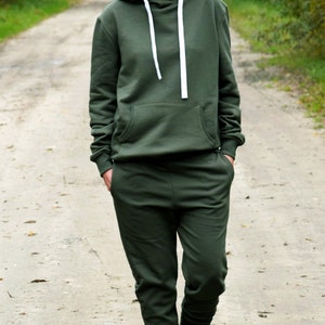 Pu&ograve; includere: Una donna indossa una felpa con cappuccio verde scuro e pantaloni della tuta abbinati. &Egrave; in piedi su un sentiero sterrato con alberi su entrambi i lati. Indossa scarpe da ginnastica bianche.