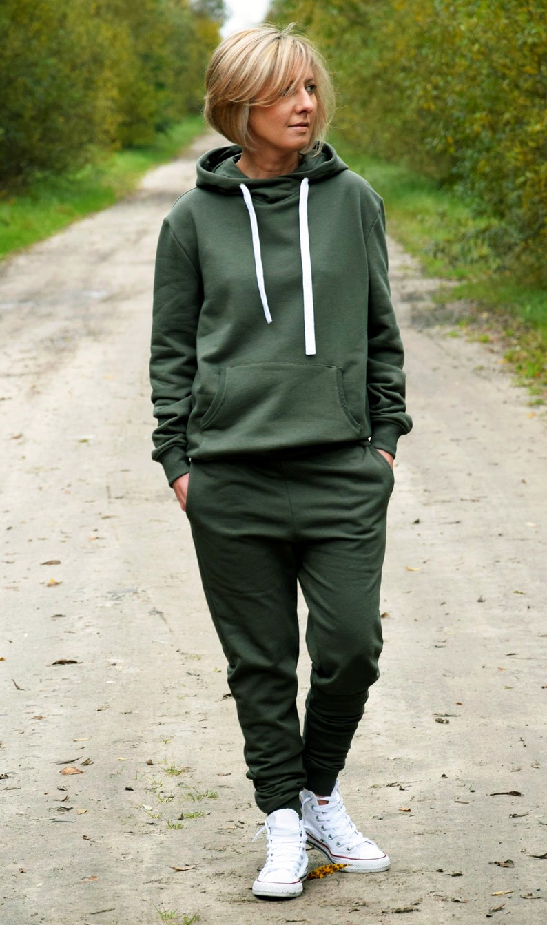Pu&ograve; includere: Una donna indossa una felpa con cappuccio verde scuro e pantaloni della tuta abbinati. &Egrave; in piedi su un sentiero sterrato con alberi su entrambi i lati. Indossa scarpe da ginnastica bianche.