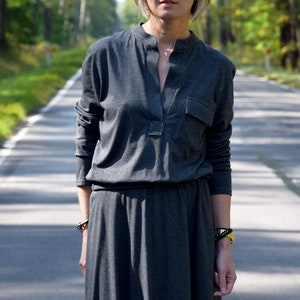 May include: A woman wearing a gray long-sleeved dress with a v-neck and a pocket on the left side. She is wearing sunglasses and a silver necklace.