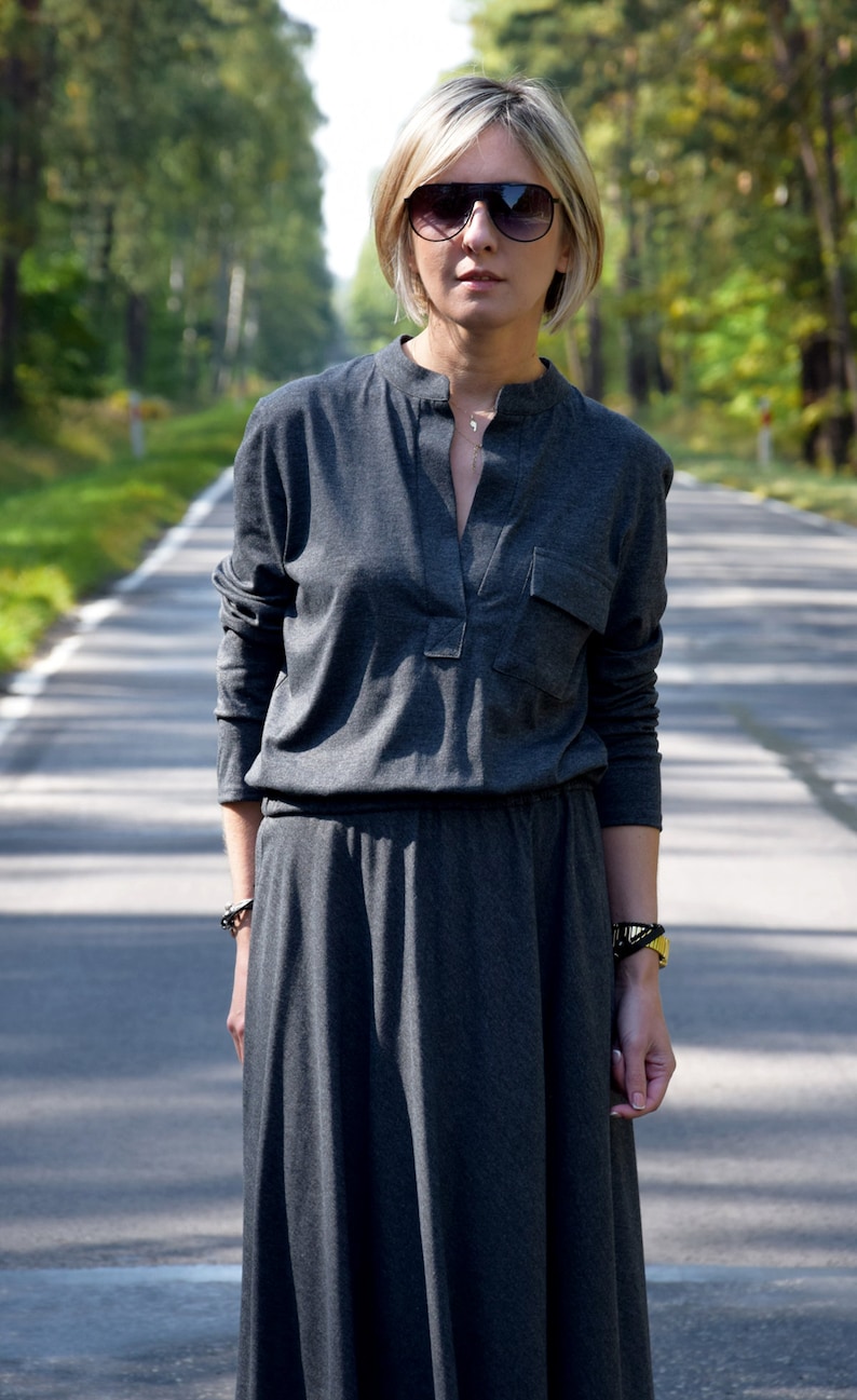 May include: A woman wearing a gray long-sleeved dress with a v-neck and a pocket on the left side. She is wearing sunglasses and a silver necklace.