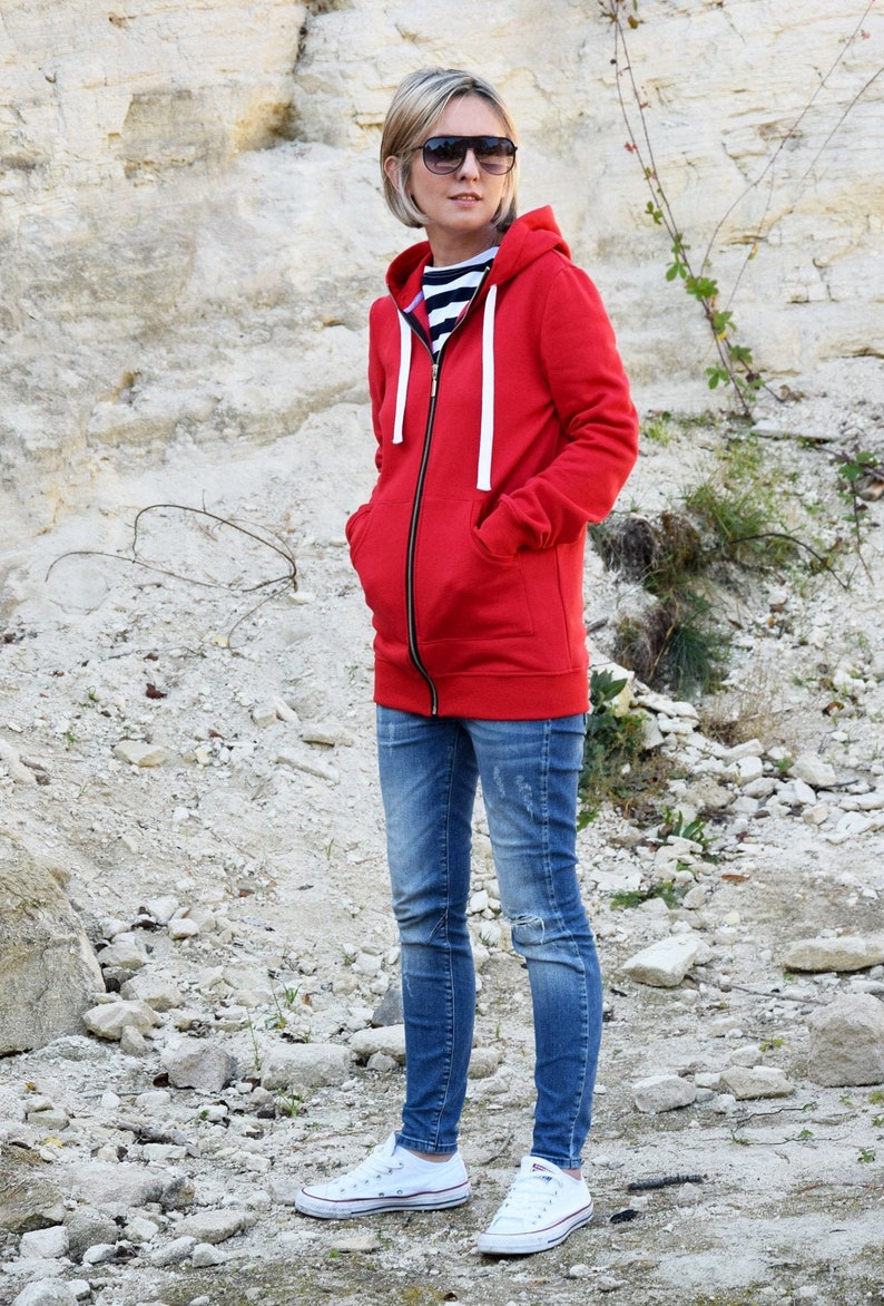 Pode incluir: Uma mulher vestindo um moletom vermelho com z&iacute;per e cord&otilde;es brancos, jeans azul e t&ecirc;nis branco. Ela est&aacute; em p&eacute; em frente a um fundo rochoso.