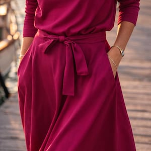 May include: A long-sleeved, burgundy dress with a matching tie belt. The dress features a round neckline and pockets. The model is wearing a watch and bracelet. The dress is made of a soft, flowing fabric.