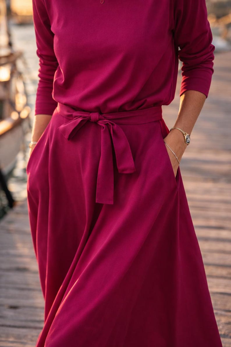May include: A long-sleeved, burgundy dress with a matching tie belt. The dress features a round neckline and pockets. The model is wearing a watch and bracelet. The dress is made of a soft, flowing fabric.