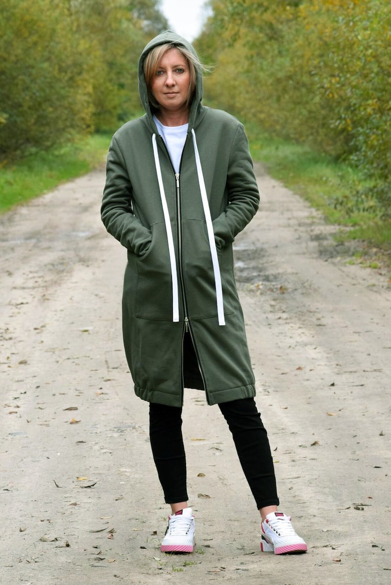 May include: A woman wearing a long, olive green hooded sweatshirt with white drawstrings and a zipper. She is standing on a gravel path with trees on either side. She is wearing black leggings and white sneakers with pink accents.