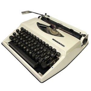 Op de afbeelding: Crèmekleurige vintage schrijfmachine met zwarte toetsen en een chromen wagenterugloophendel. De schrijfmachine heeft de merknaam "Tippa" op de voorkant gedrukt. De toetsen zijn in een QWERTY-indeling geplaatst.