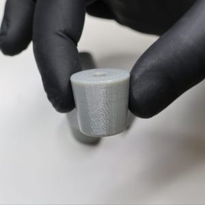 May include: A cylindrical, light gray object with a hole in the center. The object appears to be made of a solid material and has a textured surface. The object is held by a person wearing black gloves.