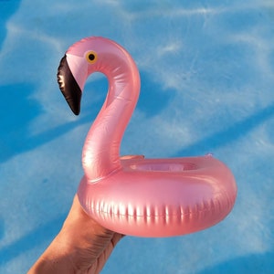 K&ouml;nnte beinhalten: Ein aufblasbarer Getr&auml;nkehalter in Form eines rosa Flamingos mit schwarzem Schnabel und gelbem Auge, vor blauem Wasser. Der Flamingo ist zum Schwimmen im Pool und zum Halten eines Getr&auml;nks konzipiert.