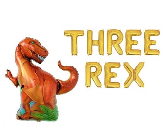 May include: A large orange and green Tyrannosaurus Rex shaped foil balloon positioned next to gold foil letter balloons that spell out the phrase THREE REX. The dinosaur balloon features a detailed reptilian skin pattern and a wide open mouth.