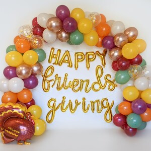 May include: An arch of balloons in shades of orange, yellow, green, white, purple, and metallic gold, featuring gold foil letter balloons that spell out "HAPPY friends giving" and a large turkey-shaped foil balloon at the bottom left corner.