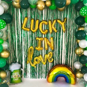 Könnte beinhalten: Grüne, goldene und weiße Ballons mit Kleeblatt- und Tupfenmustern sind um eine grüne und weiße Girlanden-Kulisse angeordnet. Die Kulisse hat ein goldenes Folien-Schild mit der Aufschrift "Lucky in Love". Ein goldener Folien-Regenbogenballon befindet sich in der rechten unteren Ecke. Ein grüner und weißer Bierkrug-Ballon mit dem Text "Happy St. Patrick's Day!" befindet sich in der linken unteren Ecke.