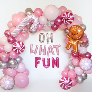 May include: Pink, silver, and brown balloon arch with candy cane, gingerbread man, and swirl designs. The arch spells out "Oh What Fun" in pink and silver letters.