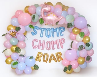 May include: A pastel-coloured balloon arch with a pink dinosaur balloon wearing a crown. The arch is made up of pink, gold, blue, and green balloons, with the words "STOMP CHOMP ROAR" in balloon letters. Green leaf accents are also included.