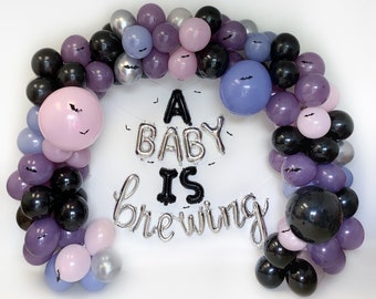 Op de afbeelding: Een paarse, zwarte en zilveren ballonboog met de tekst "A Baby Is Brewing" in zilveren en zwarte letters.