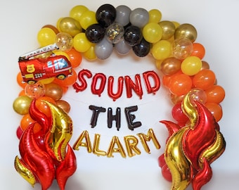 Puede incluir: Un arco de globos festivo en tonos rojos, naranjas, amarillos, dorados, grises y negros. Un globo de camión de bomberos rojo está a la izquierda. Las palabras "SOUND THE ALARM" están escritas con letras de globo.