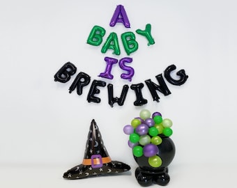 Pode incluir: Uma decoração de chá de bebê com balões pretos, verdes e roxos que soletram "A Baby Is Brewing". Também está incluído um chapéu de bruxa e um caldeirão cheio de balões verdes e roxos.