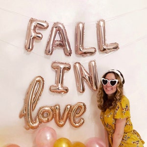 May include: A woman wearing a yellow floral dress and white heart-shaped sunglasses stands in front of a white wall with rose gold foil balloons that spell "Fall in love".  The balloons are strung together with white string.  There are pink, gold, and white polka dot balloons at the bottom of the image.