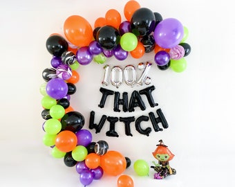 May include: A balloon arch featuring orange, black, purple, and lime green balloons arranged in a crescent shape. Silver foil balloons spell 100%, while black foil balloons spell THAT WITCH. A small witch character foil balloon sits below.