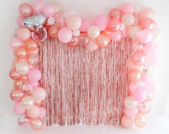 May include: A rose gold backdrop with a garland of pink and rose gold balloons. The balloons are arranged in a semi-circle shape and are tied together with clear string. The backdrop is made of rose gold foil fringe.