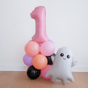 May include: A balloon arrangement featuring a large pink foil number one balloon atop a cluster of round latex balloons in shades of peach, light pink, lavender, and black, accompanied by a separate silver foil ghost balloon with a smiling face.