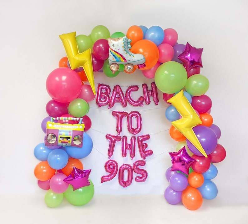 Pu&ograve; includere: Un arco di palloncini colorati con la scritta "BACH TO THE 90S". L'arco include palloncini a forma di pattino, boombox e fulmine. I palloncini sono rosa, verdi, arancioni, viola e blu, creando un'atmosfera festosa.