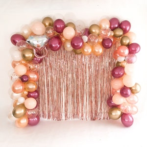 May include: A decorative balloon arch featuring a mix of metallic gold, rose gold, and burgundy balloons, accented with clear confetti-filled balloons and a large silver and pink diamond ring-shaped foil balloon over a rose gold fringe curtain.