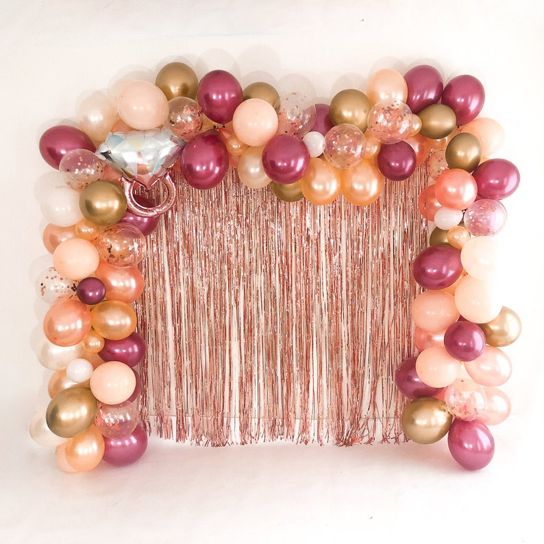 May include: A decorative balloon arch featuring a mix of metallic gold, rose gold, and burgundy balloons, accented with clear confetti-filled balloons and a large silver and pink diamond ring-shaped foil balloon over a rose gold fringe curtain.
