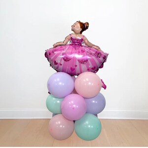 May include: A festive balloon arrangement featuring a ballerina-shaped foil balloon in a pink dress, atop a stack of pastel-colored round balloons. The balloons are in shades of pink, purple, and mint green, creating a cheerful and celebratory display.