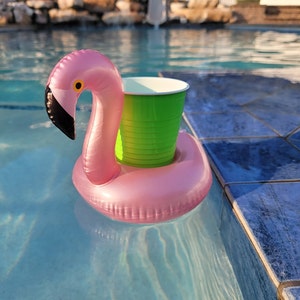 K&ouml;nnte beinhalten: Rosa aufblasbarer Flamingo-Poolschwimmer mit einem gr&uuml;nen Becherhalter, der in einem blauen Schwimmbecken schwimmt. Der Flamingo hat einen schwarzen Schnabel und ein gelbes Auge. Der Pool hat blaue Fliesen.