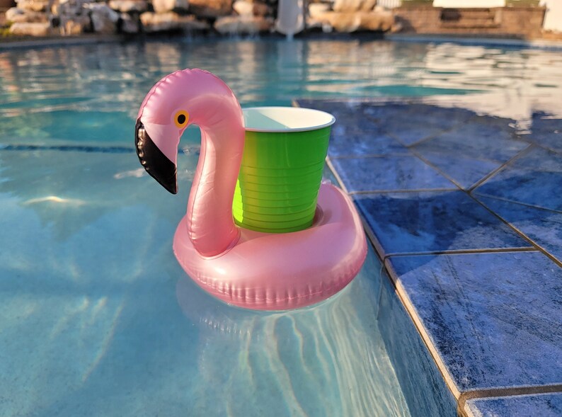 K&ouml;nnte beinhalten: Rosa aufblasbarer Flamingo-Poolschwimmer mit einem gr&uuml;nen Becherhalter, der in einem blauen Schwimmbecken schwimmt. Der Flamingo hat einen schwarzen Schnabel und ein gelbes Auge. Der Pool hat blaue Fliesen.