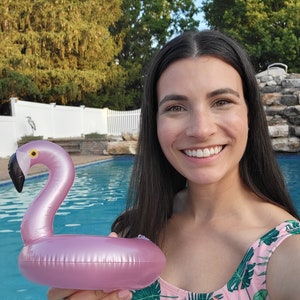 K&ouml;nnte beinhalten: Ein aufblasbarer Getr&auml;nkehalter in Form eines rosa Flamingos mit schwarzem Schnabel und gelbem Auge. Der Flamingo wird vor einem Swimmingpool gehalten. Der Hintergrund umfasst B&auml;ume und einen wei&szlig;en Zaun.