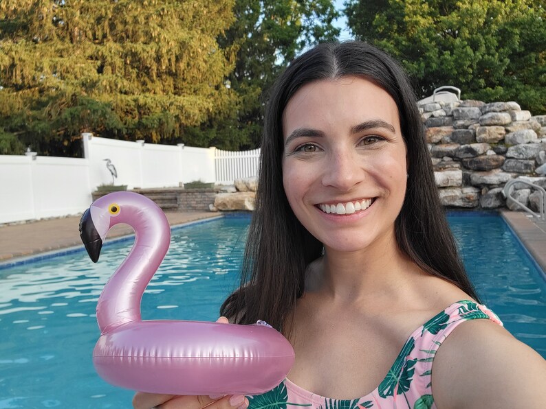 K&ouml;nnte beinhalten: Ein aufblasbarer Getr&auml;nkehalter in Form eines rosa Flamingos mit schwarzem Schnabel und gelbem Auge. Der Flamingo wird vor einem Swimmingpool gehalten. Der Hintergrund umfasst B&auml;ume und einen wei&szlig;en Zaun.