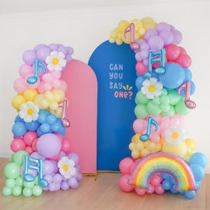 May include: A balloon arch display featuring pastel-colored balloons, white daisy-shaped balloons, metallic blue and pink musical note balloons, and a rainbow balloon. Behind the balloons are a pink arch and a blue arch with the text CAN YOU SAY ONE?