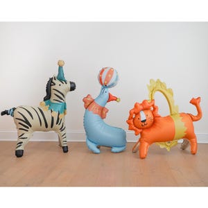 May include: Three inflatable circus animal balloons: a zebra with a party hat, a seal balancing a ball, and a lion with a yellow mane. The balloons are in a room with a light wood floor and a white wall.