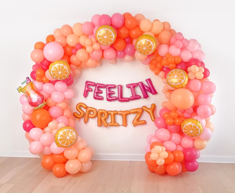 Puede incluir: Un vibrante arco de globos en tonos rosa, melocot&oacute;n y naranja, adornado con globos de rodajas de naranja. La frase "FEELIN SPRITZY" est&aacute; escrita con globos met&aacute;licos, creando un ambiente festivo y alegre.