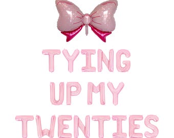 May include: Pink foil balloons spelling "TYING UP MY TWENTIES" with a pink bow-shaped balloon at the top. The bow is light pink with white polka dots and magenta accents. The balloons are arranged on a white background.