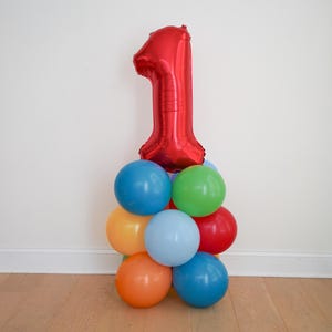 May include: A large red foil balloon shaped like the number one, positioned atop a cluster of smaller, round latex balloons in various colors including blue, green, yellow, light blue, orange, and red, creating a festive balloon arrangement.