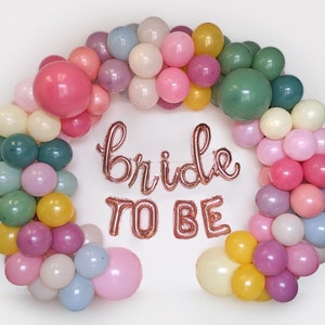 Puede incluir: Un arco de globos coloridos en tonos rosa, verde, amarillo y morado, con las palabras "bride to be" en globos de papel de oro rosa. Los globos est&aacute;n dispuestos en forma de arco, perfecto para una despedida de soltera o boda.