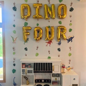 May include: Gold foil balloons spelling DINO FOUR arranged on a wall above a white toy kitchen. Vertical garlands feature green paper monstera leaves and dinosaur footprints. Small dinosaur cutouts are interspersed between the garlands.