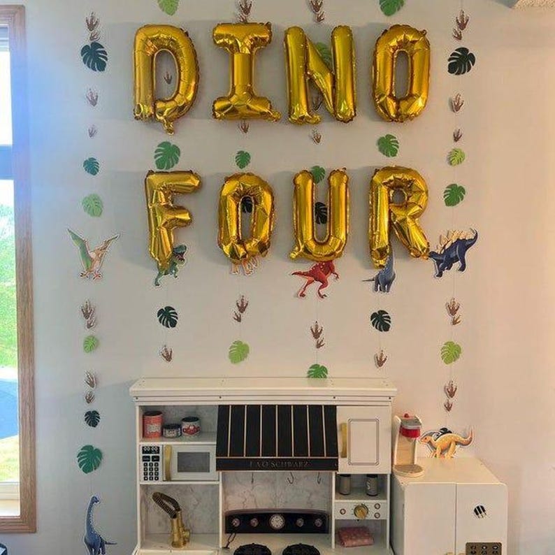 May include: Gold foil balloons spelling DINO FOUR arranged on a wall above a white toy kitchen. Vertical garlands feature green paper monstera leaves and dinosaur footprints. Small dinosaur cutouts are interspersed between the garlands.